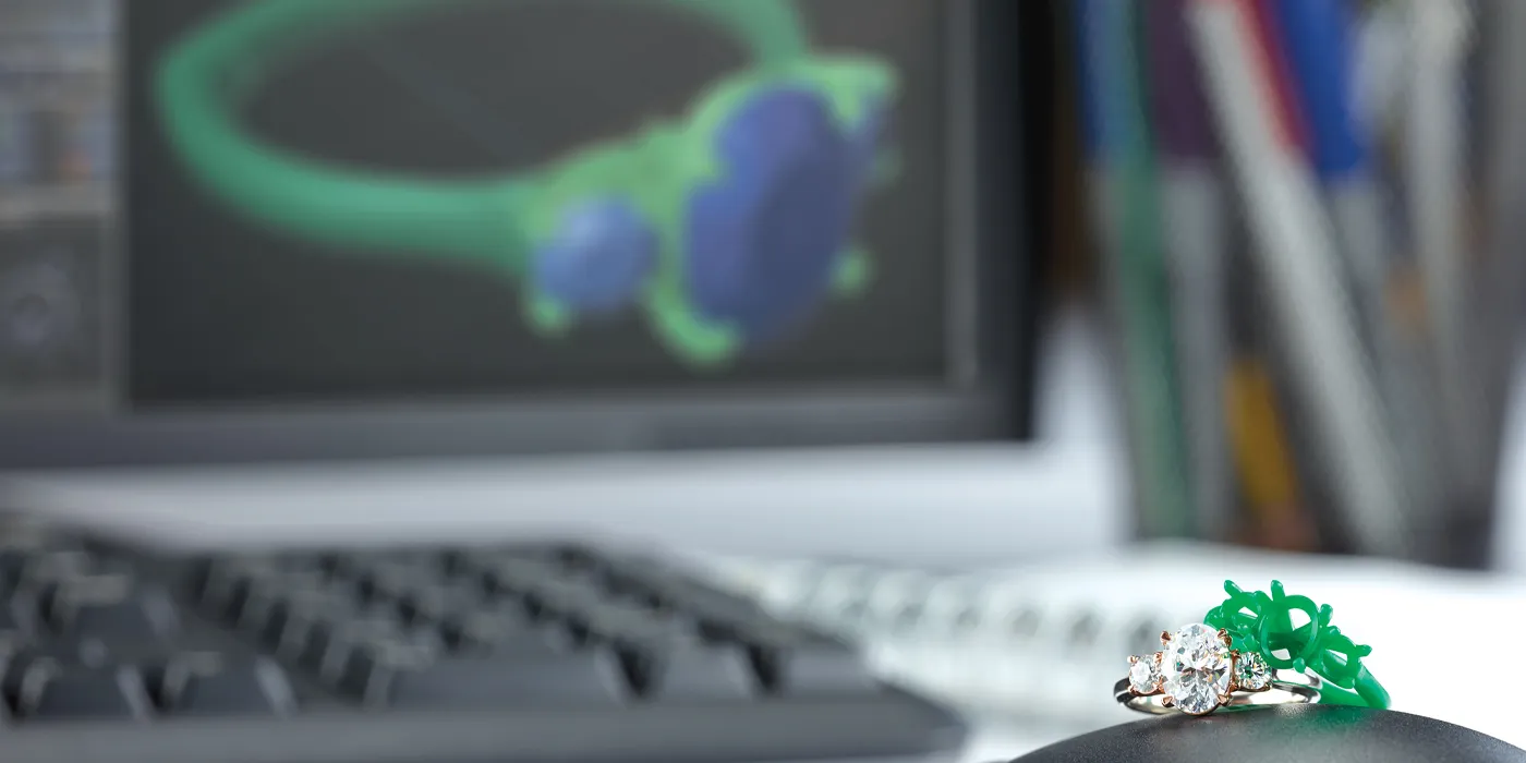 A diamond engagement ring with a green wax model sits on a computer mouse, with a design rendering in the background.