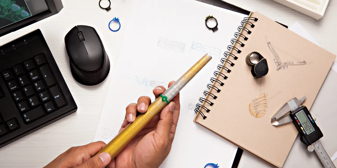 A hand holding a ring sizer tool over sketches of jewelry designs, with a keyboard and mouse nearby.
