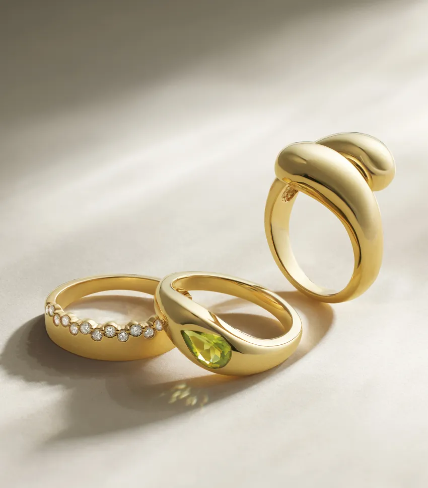 Three elegant gold rings featuring a diamond-studded band and a green gemstone, displayed on a soft surface.