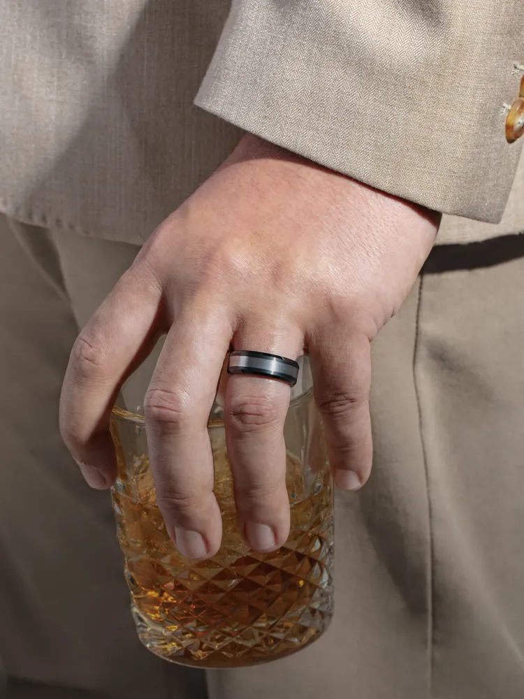 A hand holding a whiskey glass, wearing a black and silver men's fashion ring. Shop Men's Fashion Rings at Monarch Jewelry.
