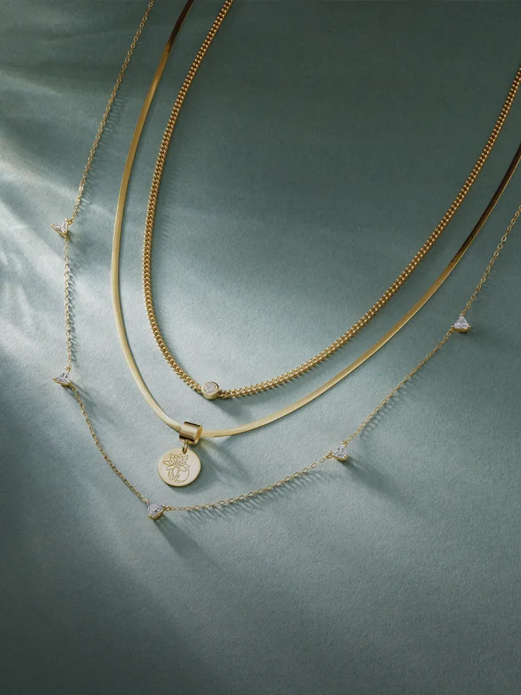 Three elegant gold necklaces with a pendant and sparkling accents, displayed on a soft green background. Shop Necklaces.