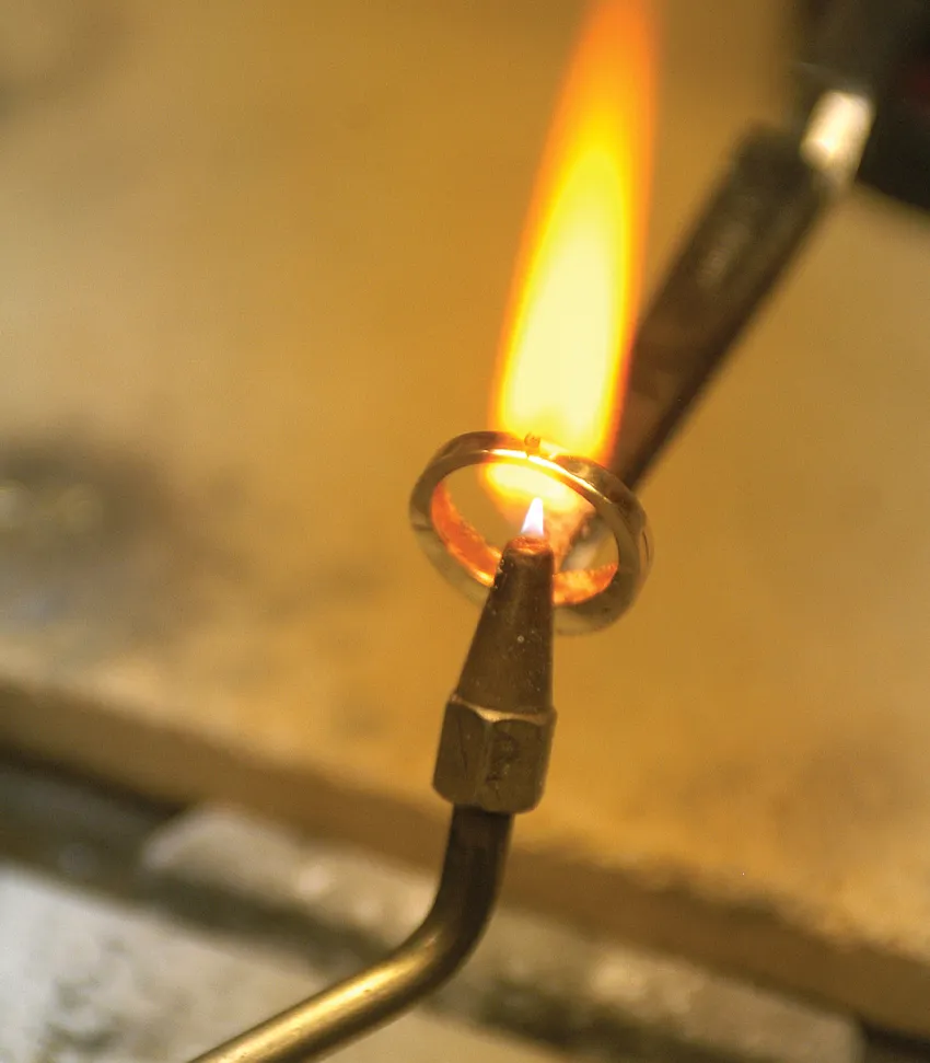 A jeweler uses a torch to heat a metal ring, creating a flame as part of the jewelry-making process.