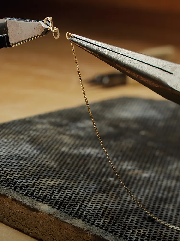 Goldsmith repairing a chain clasp with pliers on a workbench. Click to learn more about our repair services at Monarch Jewelry.