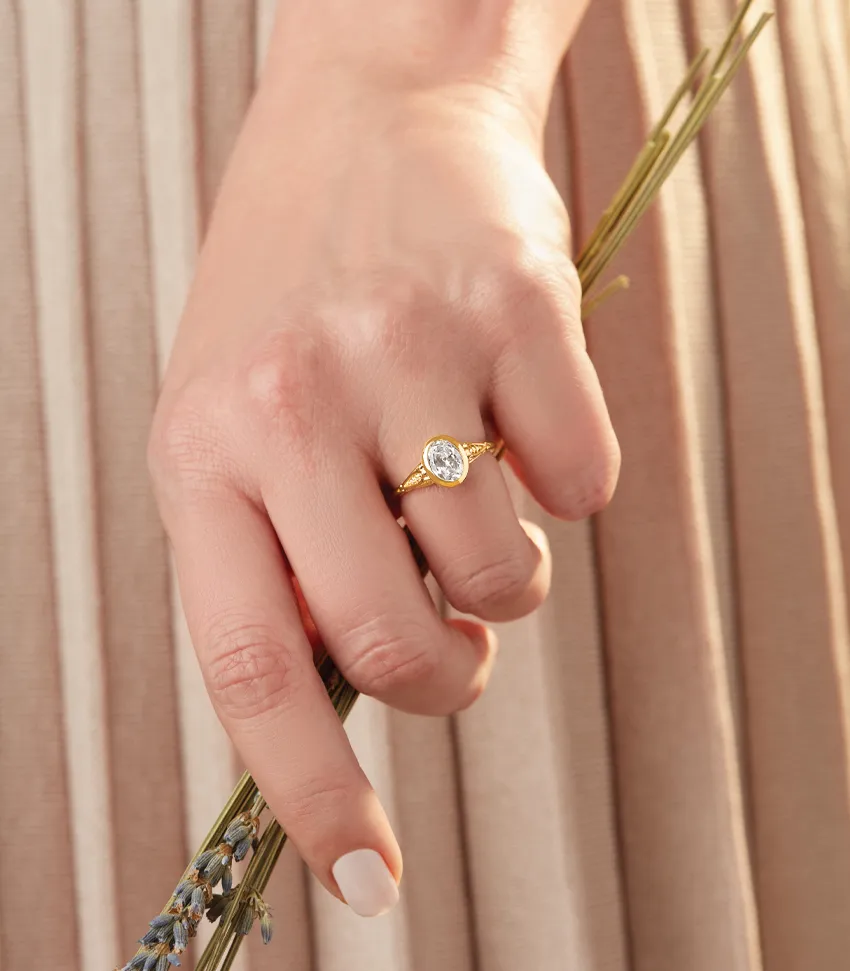 A hand holding lavender with a gold engagement ring featuring a pear-shaped diamond. Visit Monarch Jewelry for more.