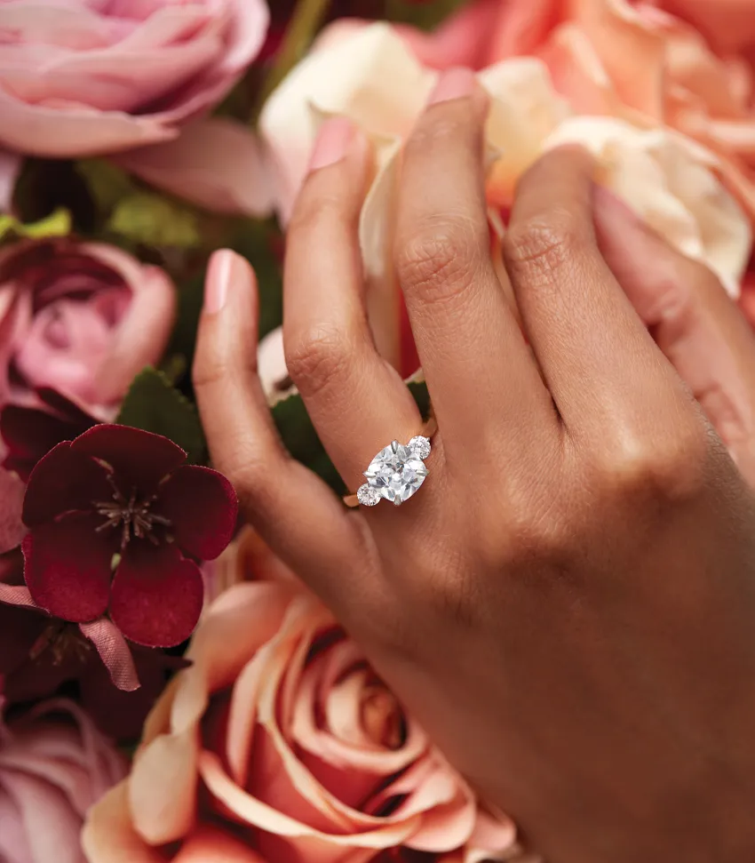 A hand wearing an elegant engagement ring with a large diamond, surrounded by colorful roses. Visit Monarch Jewelry for more.