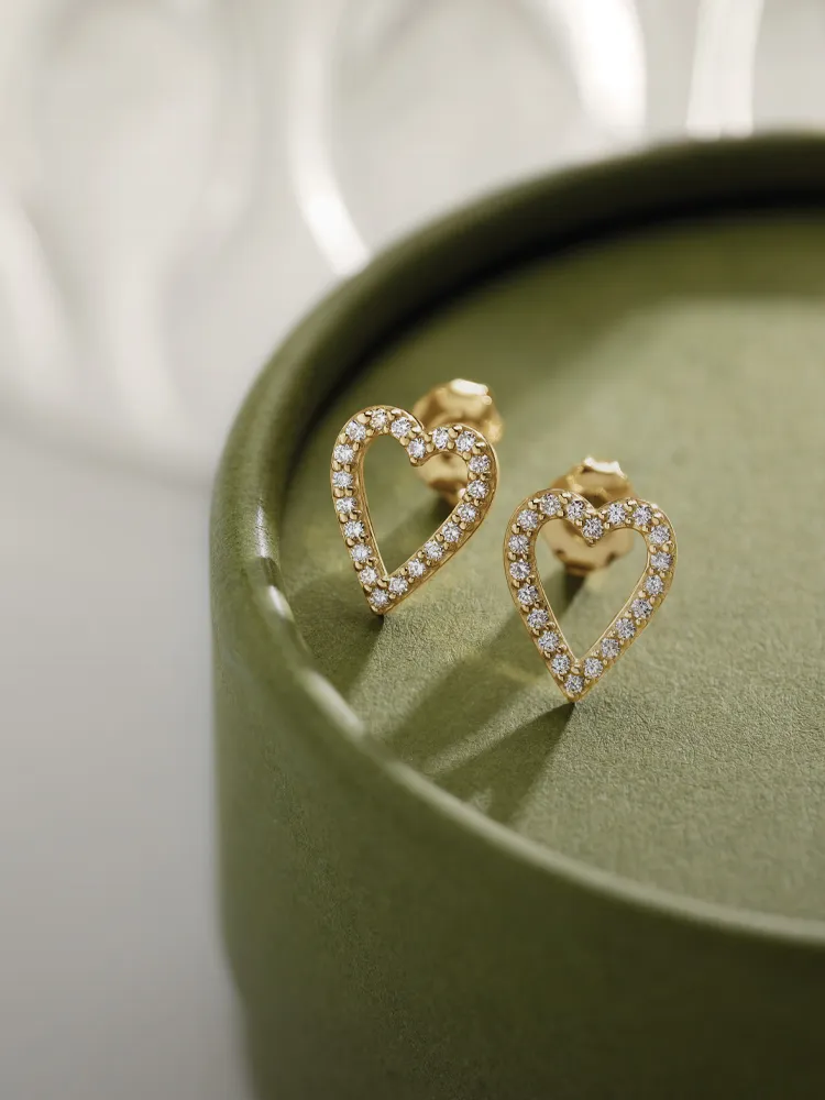 Gold heart-shaped earrings adorned with sparkling diamonds, displayed on a green box. Shop Earrings at Monarch Jewelry.