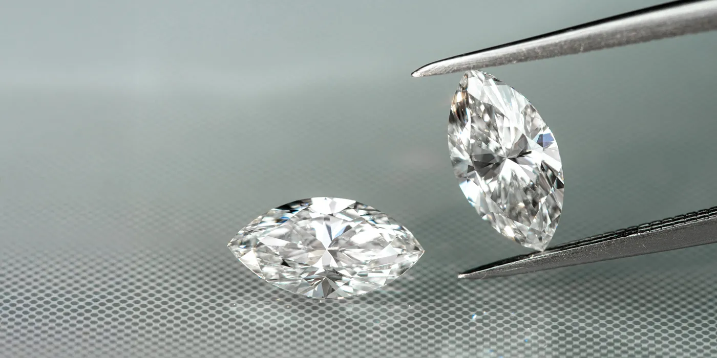 Two sparkling marquise-cut diamonds being examined with tweezers on a textured surface. Visit Monarch Jewelry for more.