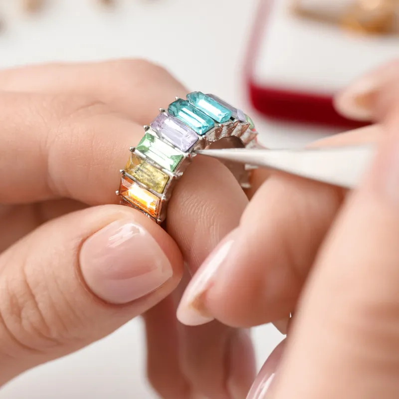 A hand holding a ring with colorful gemstones, using tweezers for adjustment. Visit Monarch Jewelry for fine jewelry.