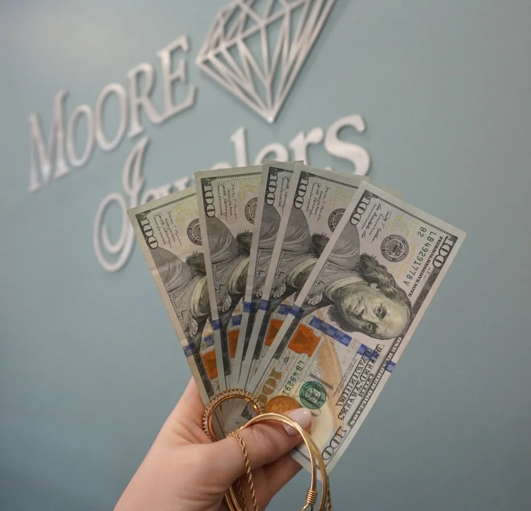 Hand holding gold jewelry and hundred dollar bills in front of Moore Jewelers sign