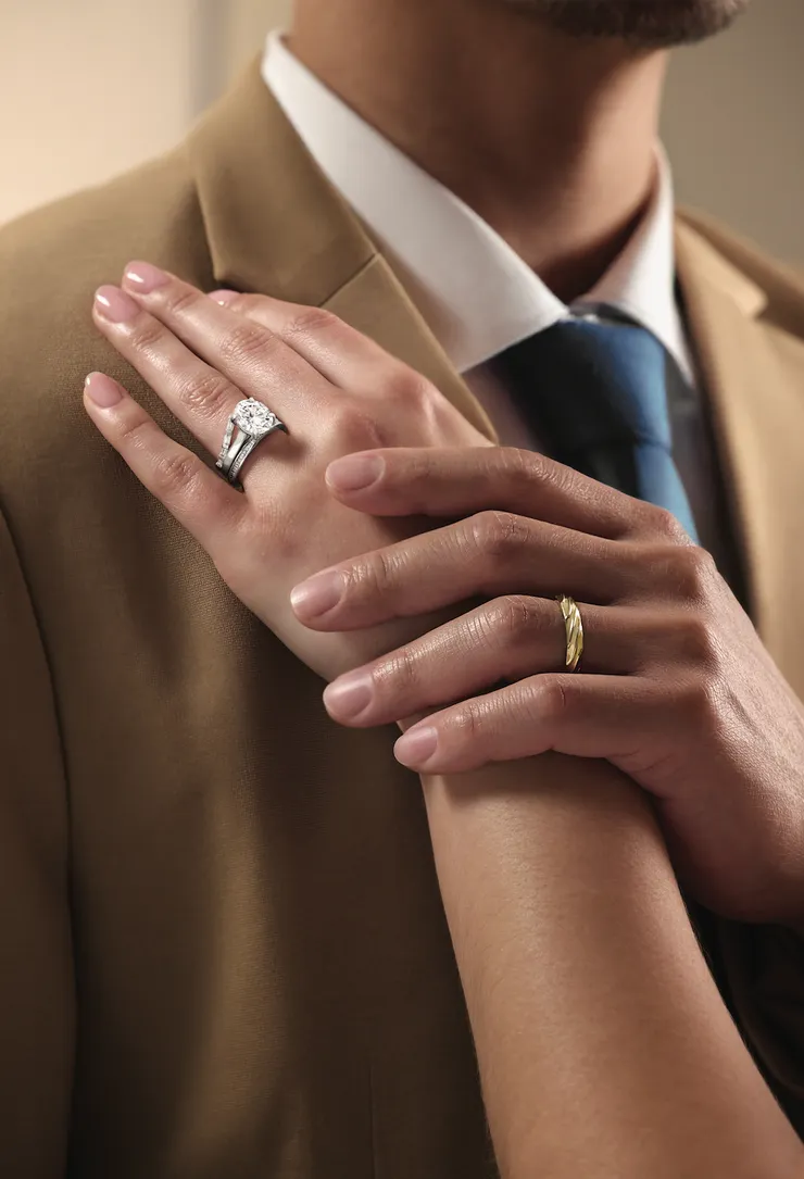 A hand wearing a diamond engagement ring with a pavé band rests on a man's shoulder, beside a hand wearing a gold wedding band