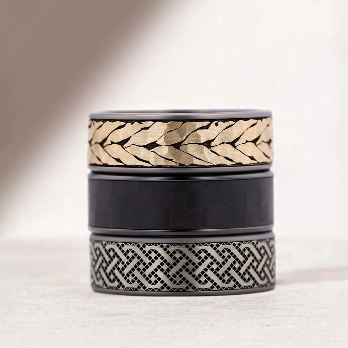 Three stacked rings with intricate designs: gold leaf pattern, plain black, and silver Celtic knot.