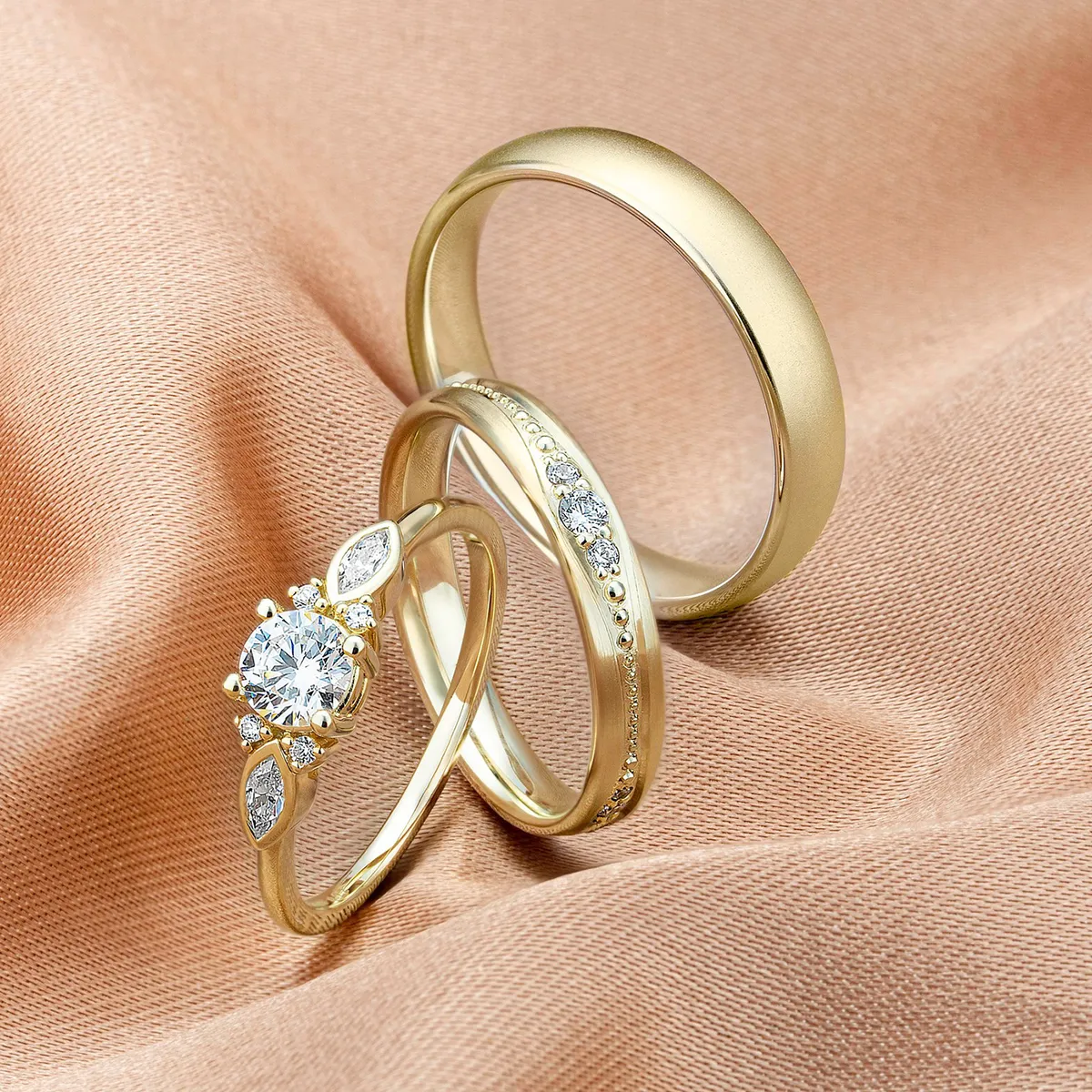 Three gold rings with diamonds on a peach fabric background.