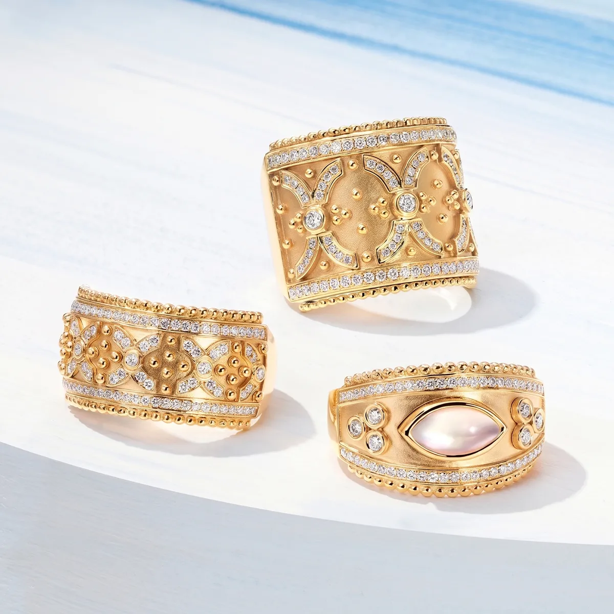 Three ornate gold rings with intricate designs and gemstones on a light surface.