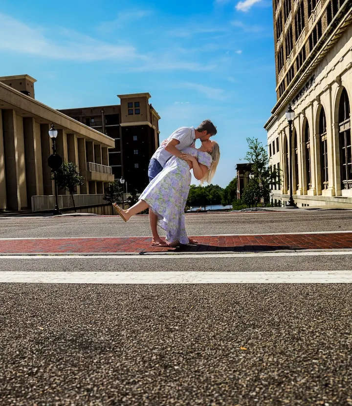 A couple shares a romantic kiss on a city street, surrounded by tall buildings.