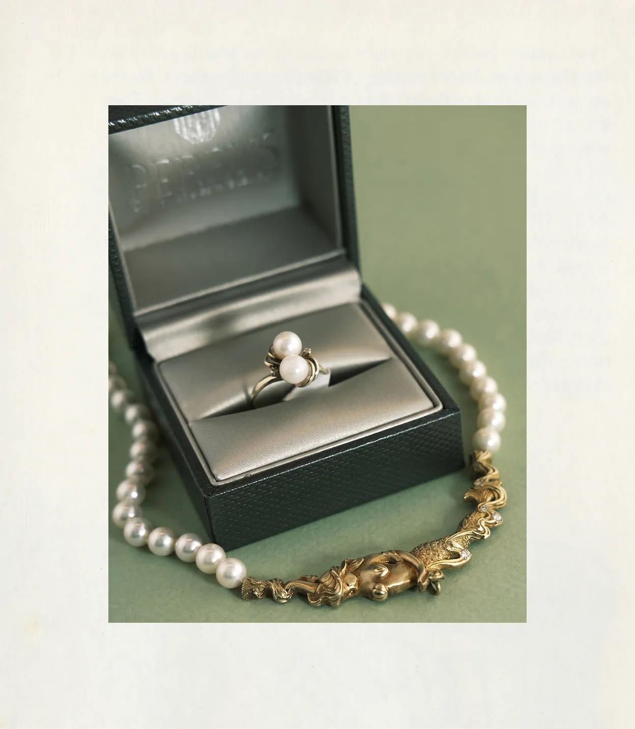 A pearl ring in a jewelry box with a pearl necklace on a green surface.
