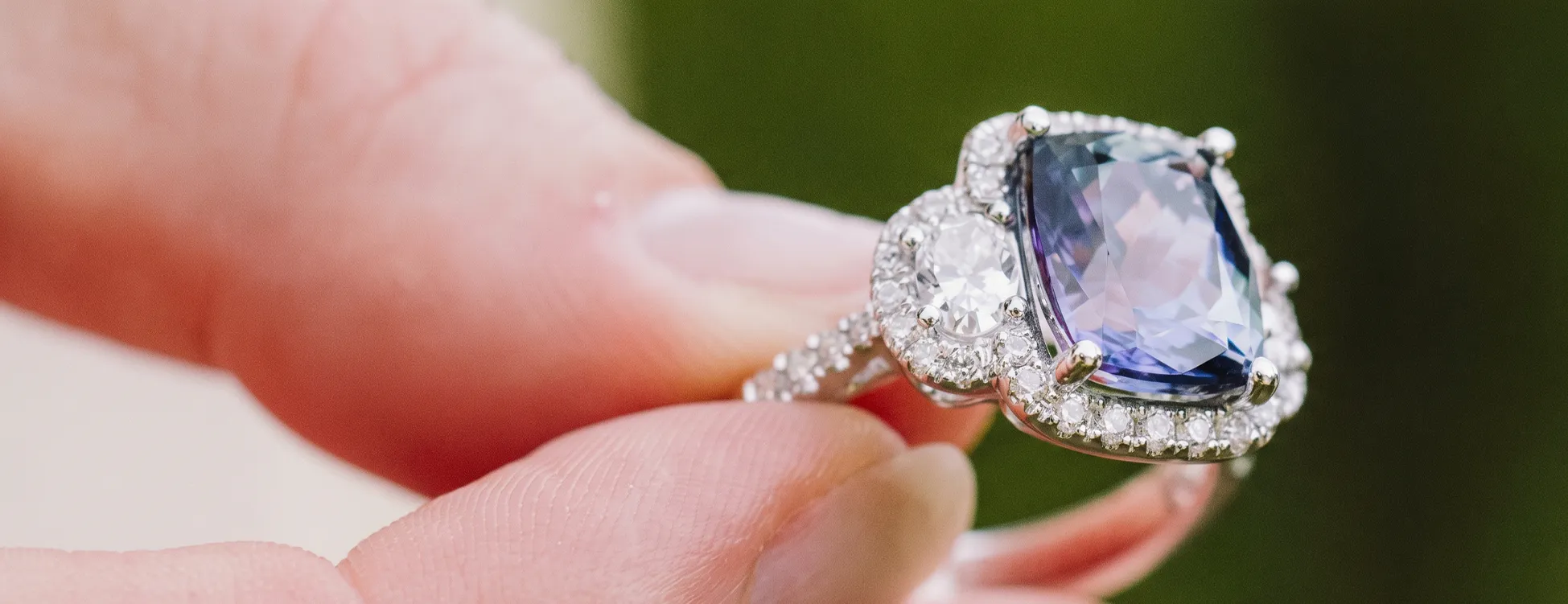 A hand holding a silver ring with a large blue gemstone and surrounding diamonds.