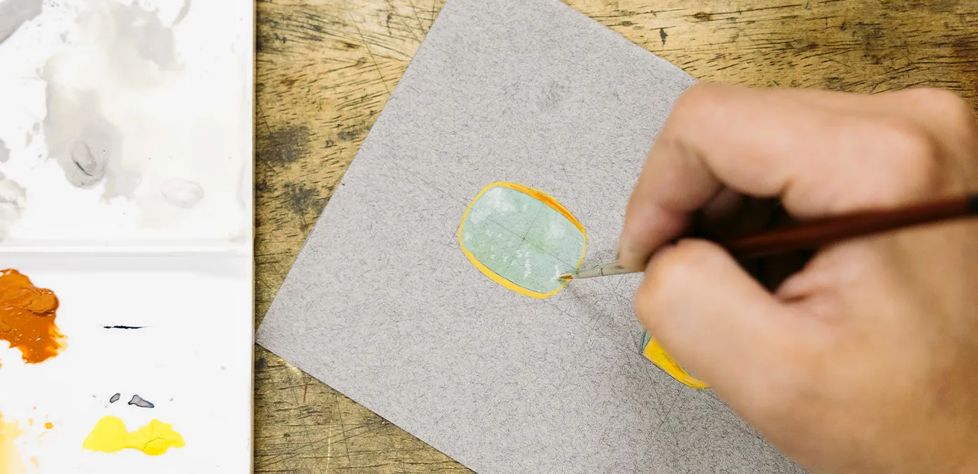 A hand painting a design on paper with a brush, next to a palette with paint.
