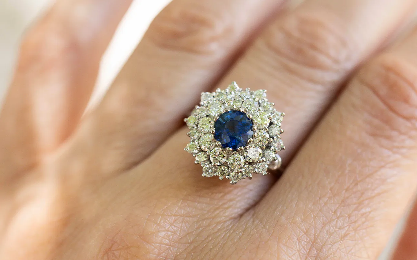 A hand wearing a ring with a blue gemstone surrounded by diamonds.