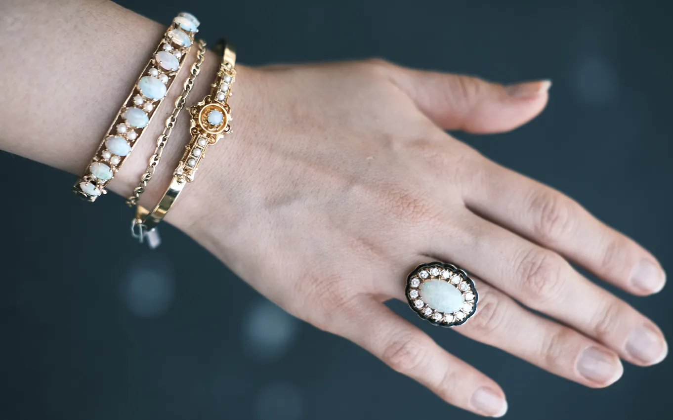 A hand wearing two gold bracelets with opal stones and an opal ring with a pearl border.