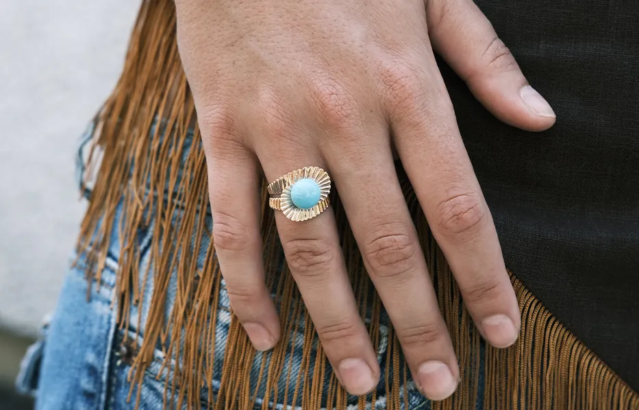 A hand wearing a gold ring with a large turquoise stone, against a fringed fabric background.