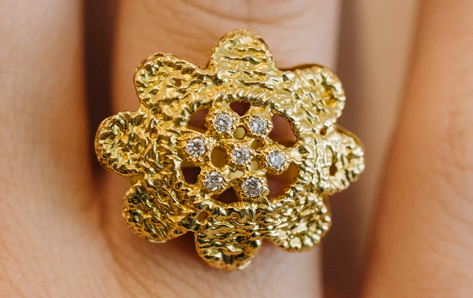 A textured gold ring with a floral design and small diamonds on a finger.
