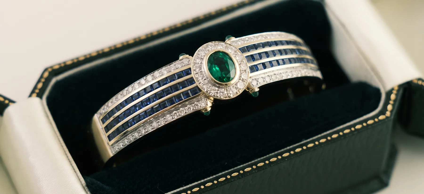 A luxurious bracelet with emerald, diamonds, and sapphires in an open jewelry box.