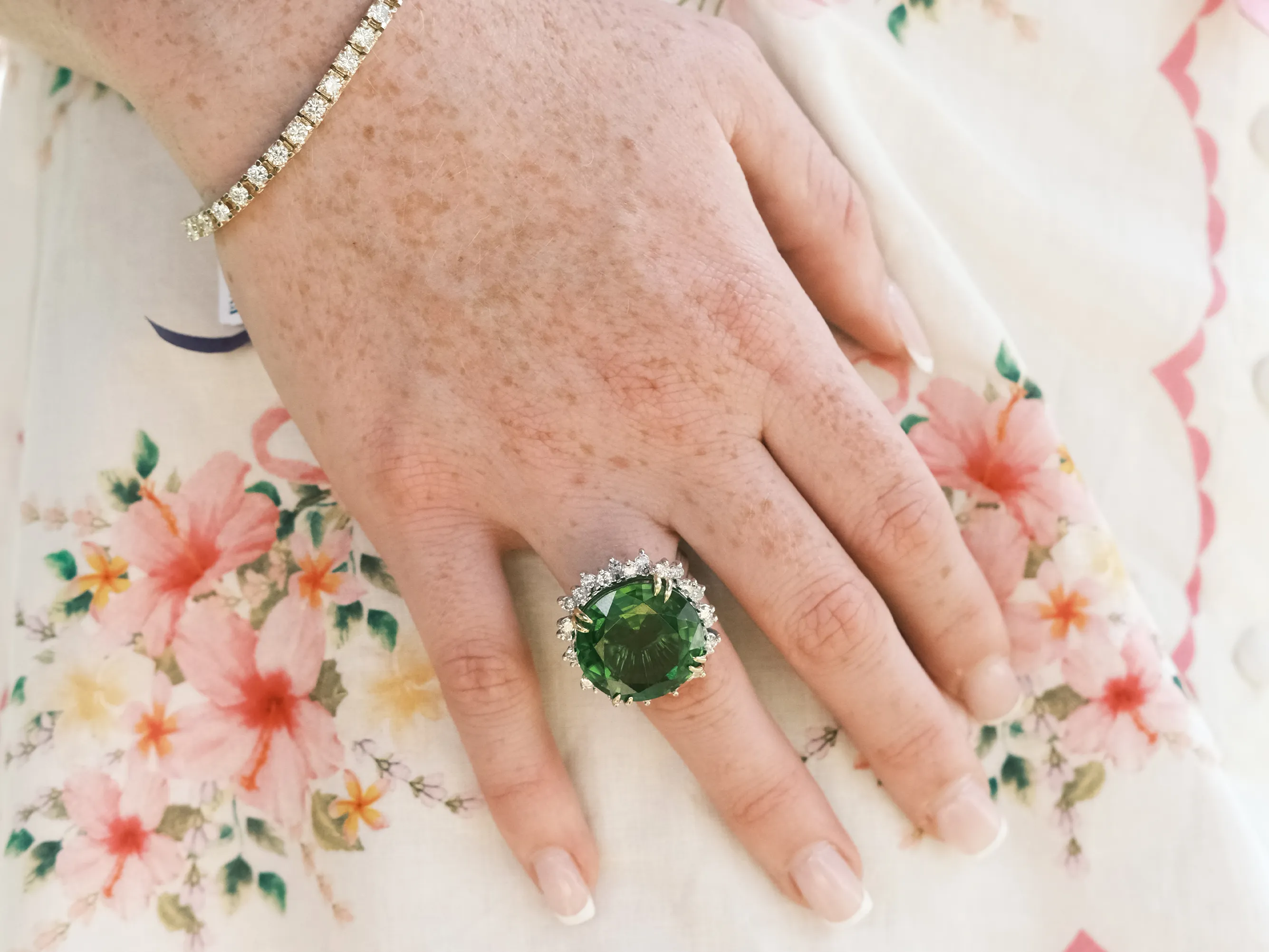 A hand with a large green gemstone ring and diamond bracelet on a floral fabric background.