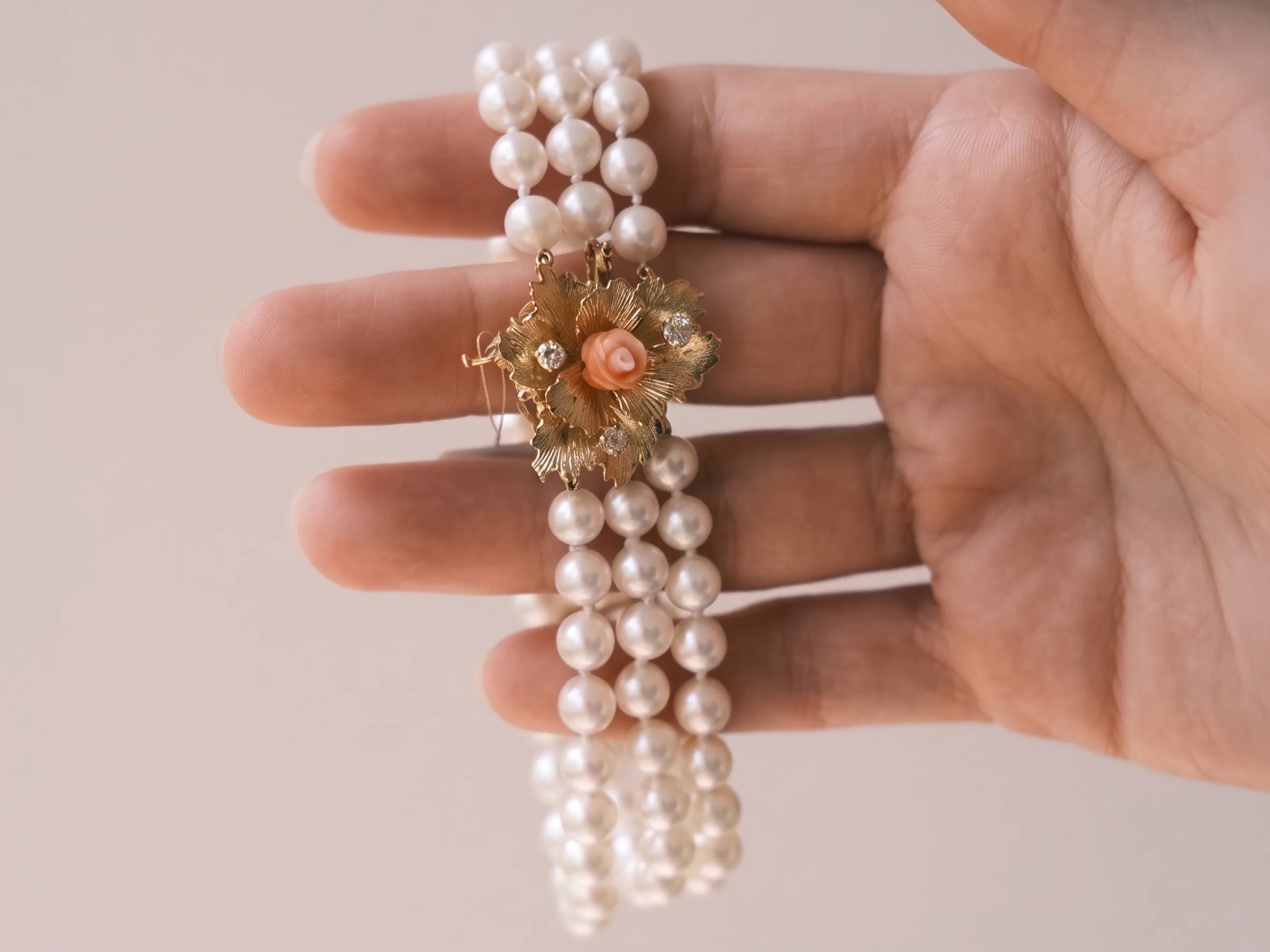 A hand holding a pearl bracelet with a floral gold clasp featuring a small pink rose.