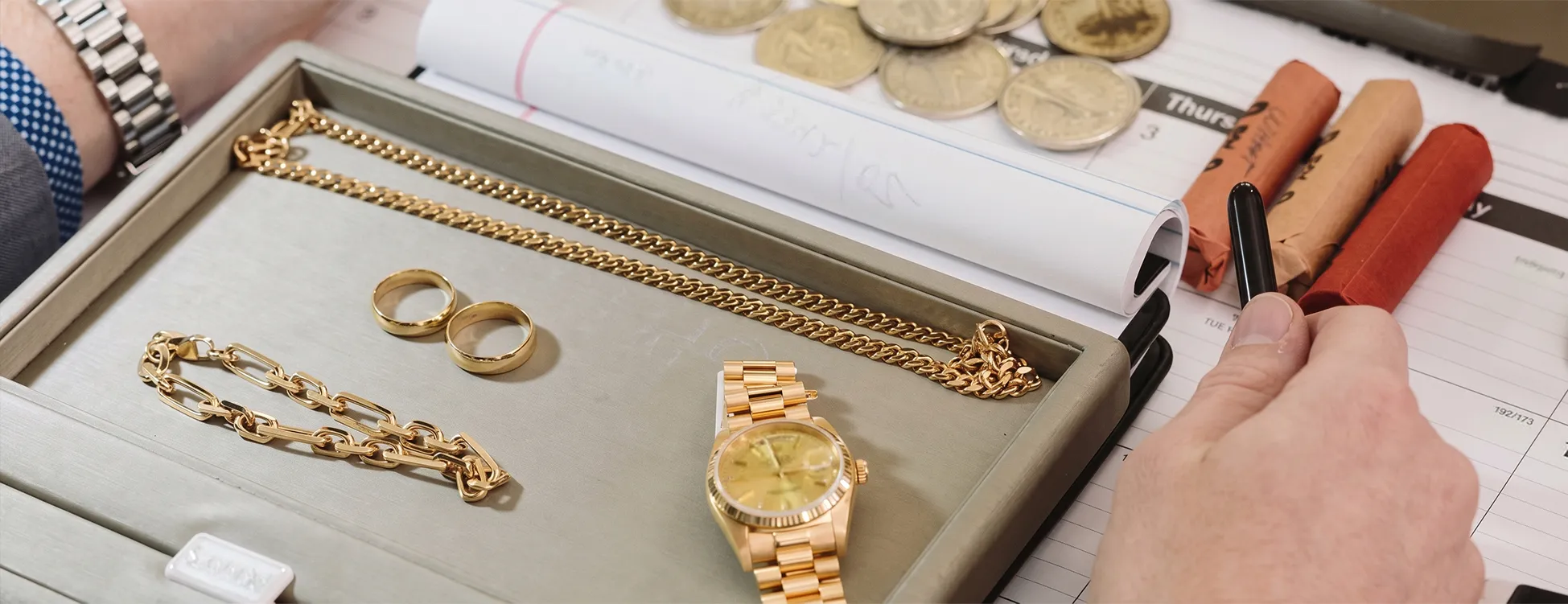 A tray with gold jewelry, including a watch, rings, and chains, next to rolled coins and a hand holding a pen.