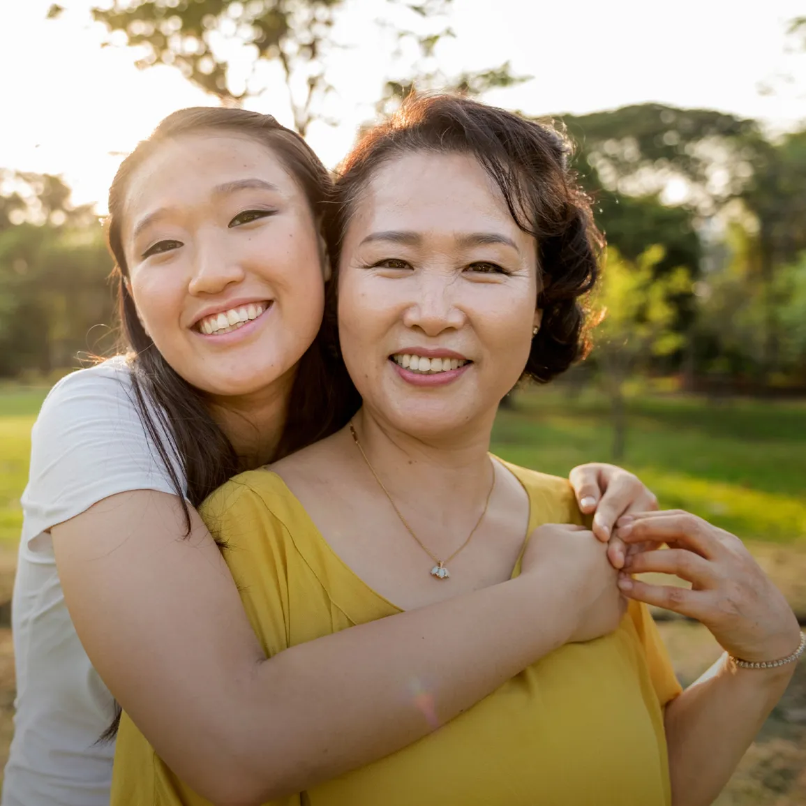 A woman in yellow and a younger woman smiling and embracing outdoors.