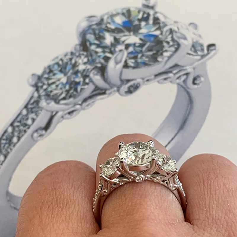 A hand wearing a three-stone diamond ring with a similar ring shown in the background.  Click for custom jewelry.