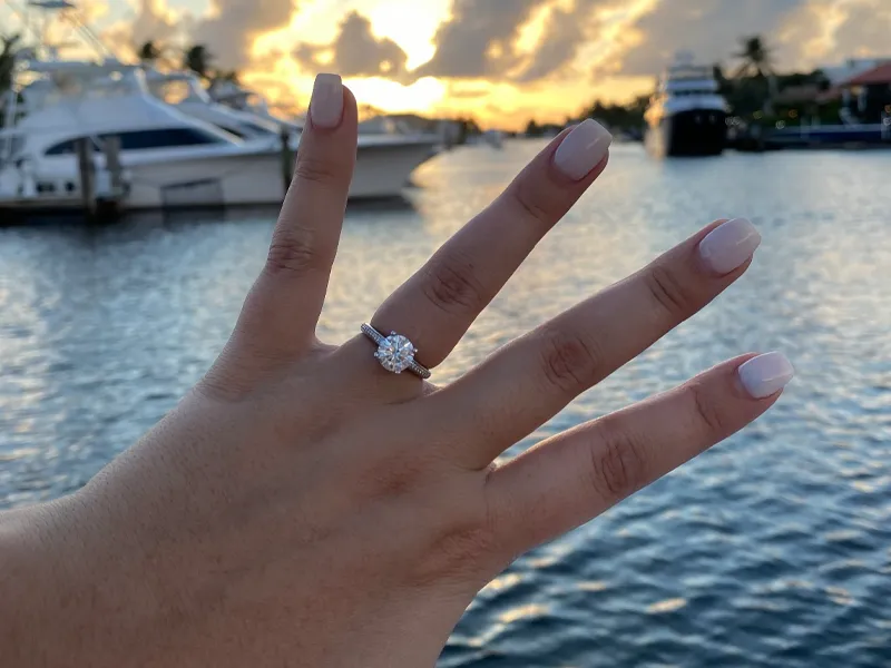 Hand displaying an engagement ring against a sunset over water, with boats in the background. Visit P.J. Rossi Jewelers.