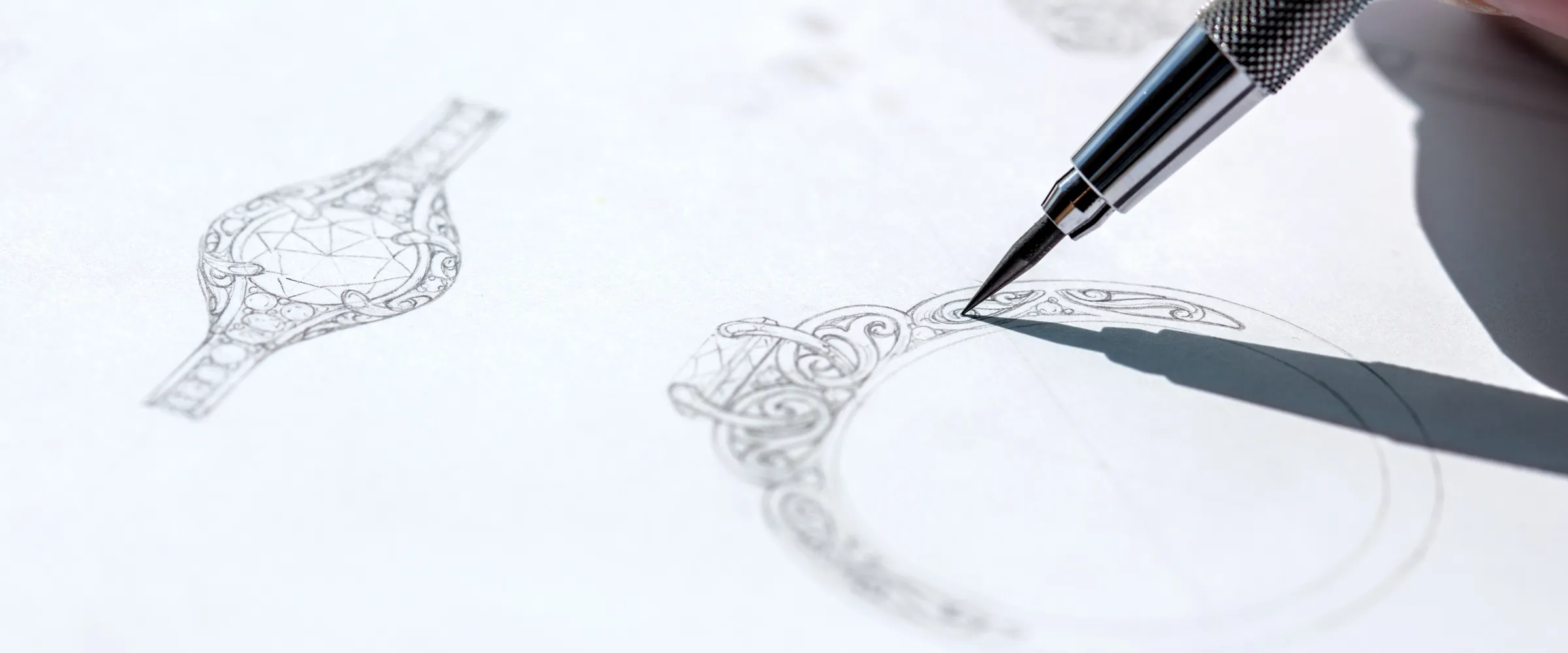 A hand sketches intricate jewelry designs on paper, showcasing custom engagement ring concepts. Click to start a project.