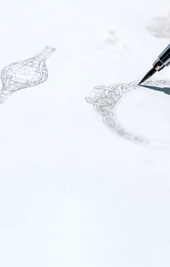 A hand sketches intricate jewelry designs on paper, showcasing custom engagement ring concepts. Click to start a project.