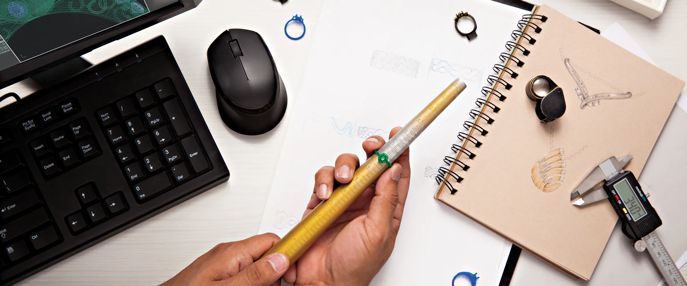 Hand holding a ring sizer over sketches and tools for jewelry design, with a computer and keyboard in the background. Learn More