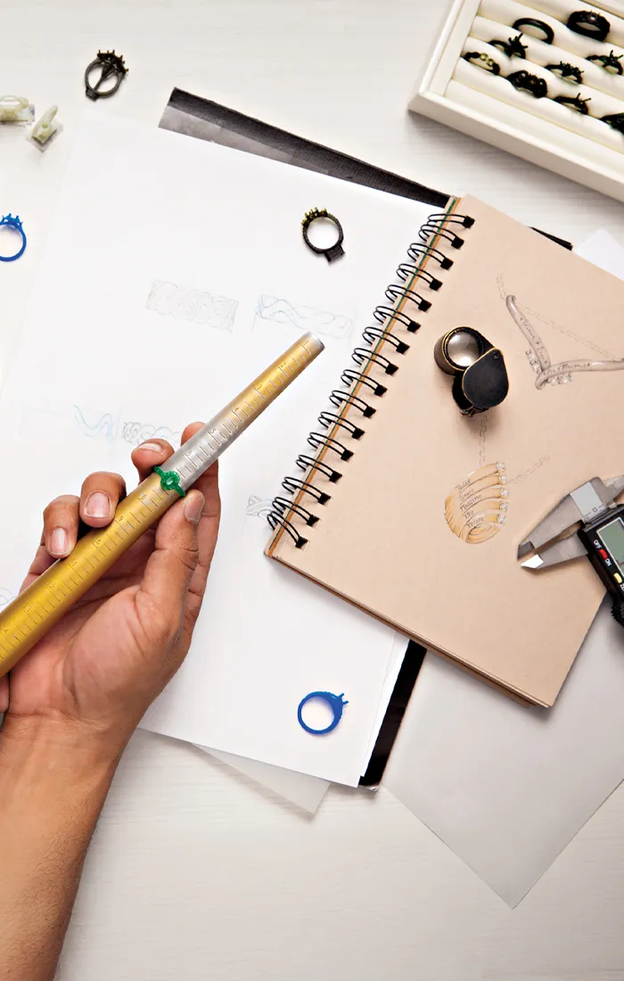 Hand holding a ring sizer over sketches and tools for jewelry design, with a computer and keyboard in the background. Learn More