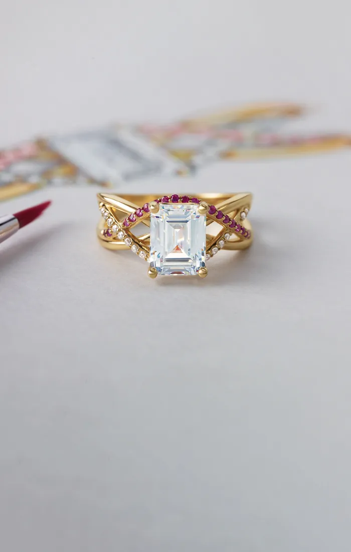 Elegant gold ring with a large emerald-cut diamond and pink gemstones, alongside a paintbrush and design sketch.
