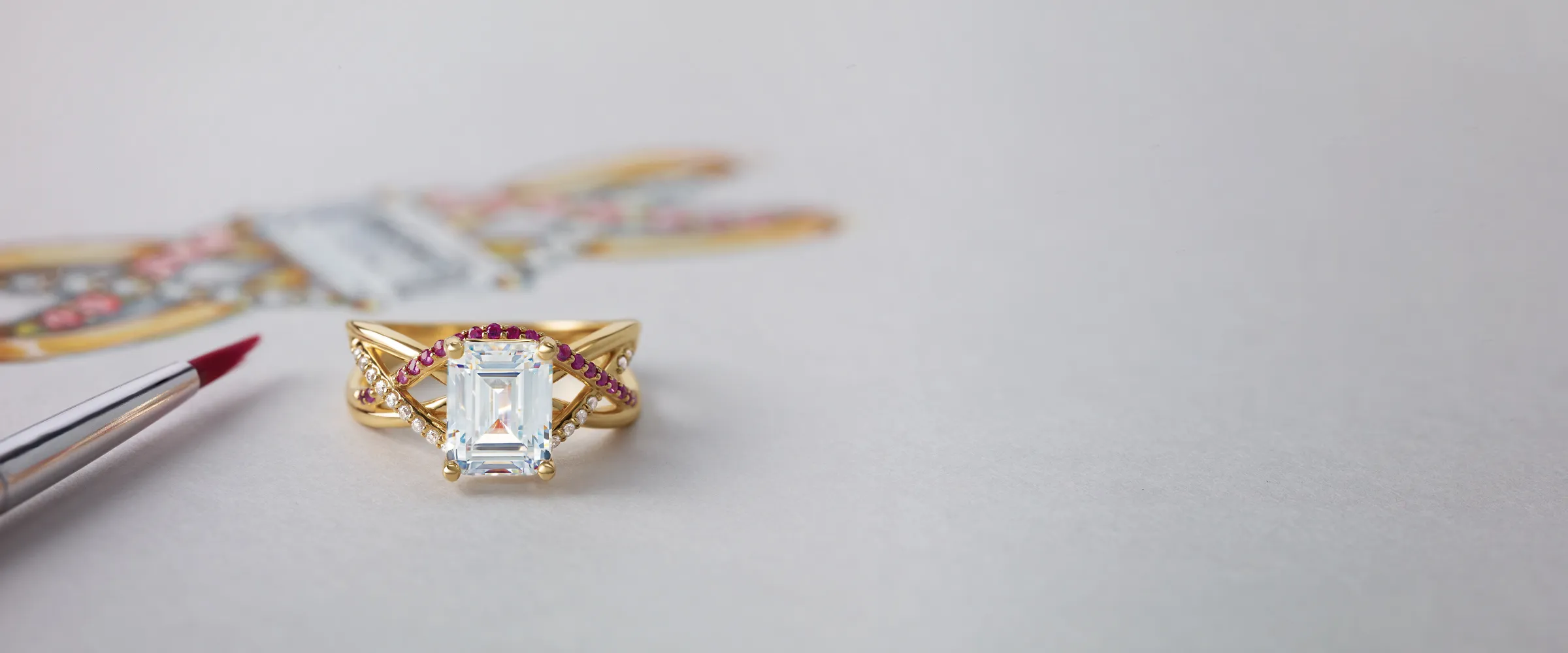 Elegant gold ring with a large emerald-cut diamond and pink gemstones, alongside a paintbrush and design sketch.