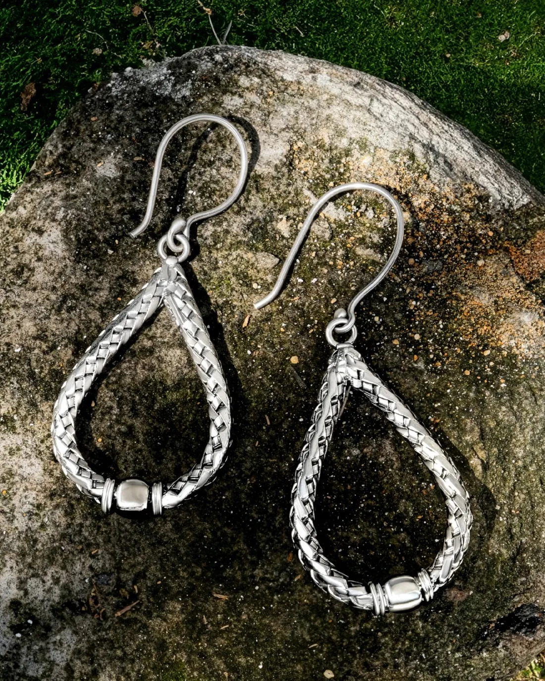 Silver teardrop earrings with a braided design, displayed on a stone.