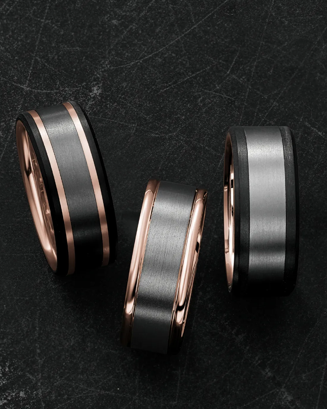 Three stylish wedding bands in black and gray with rose gold accents, displayed on a dark surface. Click to shop now.