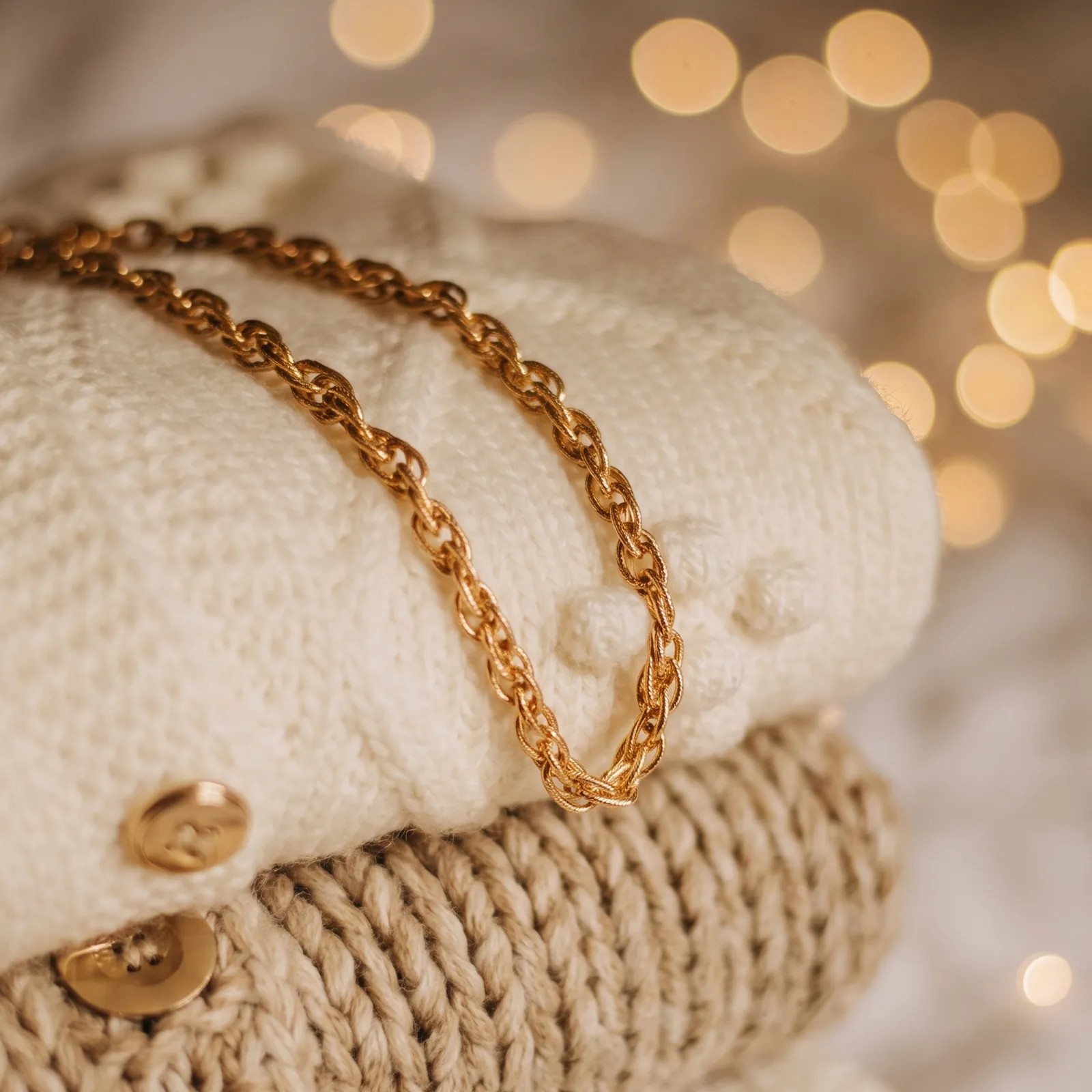 A gold chain necklace on folded knit sweaters with blurred lights in the background.