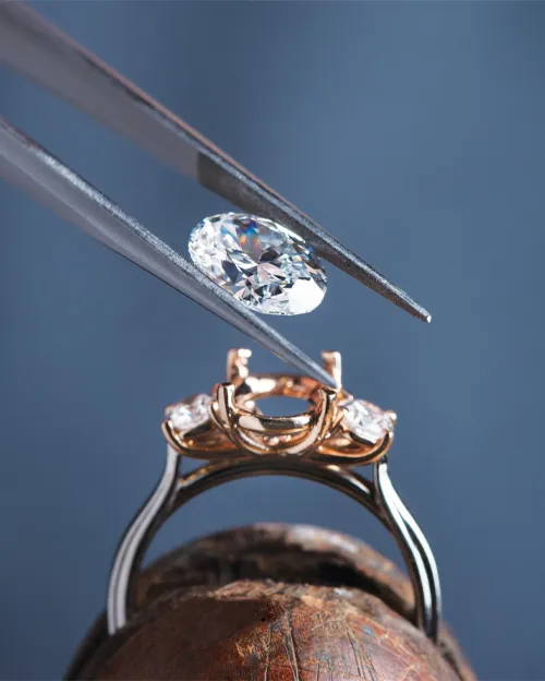 Tweezers placing a stone on ring with blue background | Jewelry Design available at Raleigh Diamond Fine Jewelry Raleigh, NC