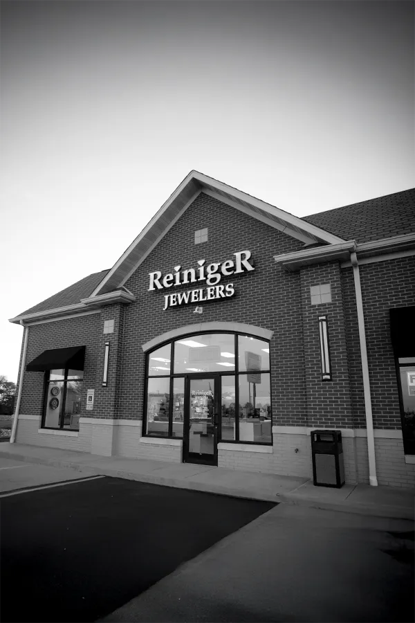 A hand sketches a detailed necklace design with a blue pencil on white paper, surrounded by other design sheets.