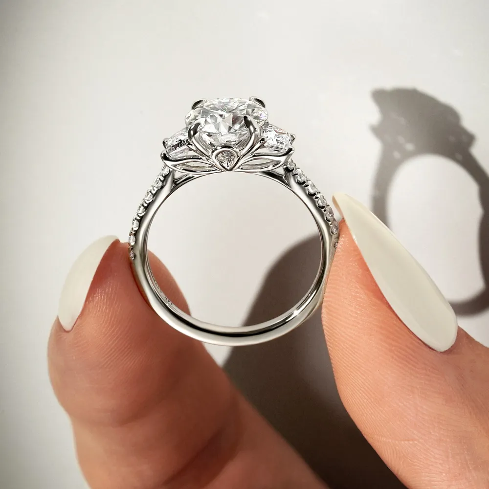 Elegant engagement ring with a round diamond centerpiece and side stones, held between fingers. Click to Shop Now.