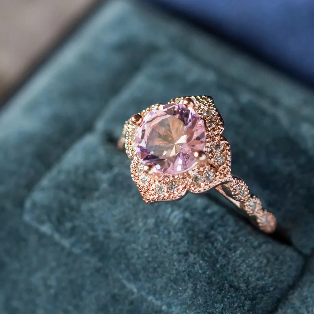 Rose gold engagement ring with a large pink gemstone surrounded by smaller diamonds, displayed on a blue velvet background. Clic