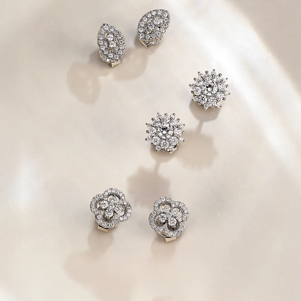 Sparkling diamond stud earrings in various floral and geometric designs, displayed on a soft, neutral background. Shop Now.