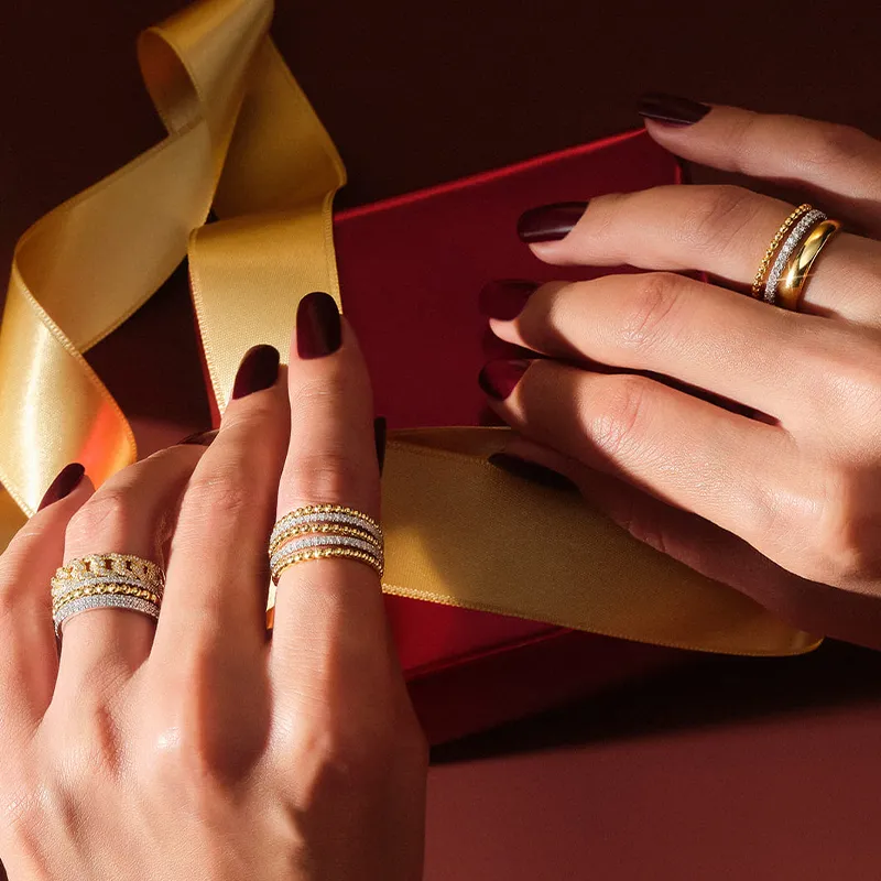 Hands with dark nails wearing multiple gold and diamond rings, holding a yellow ribbon.