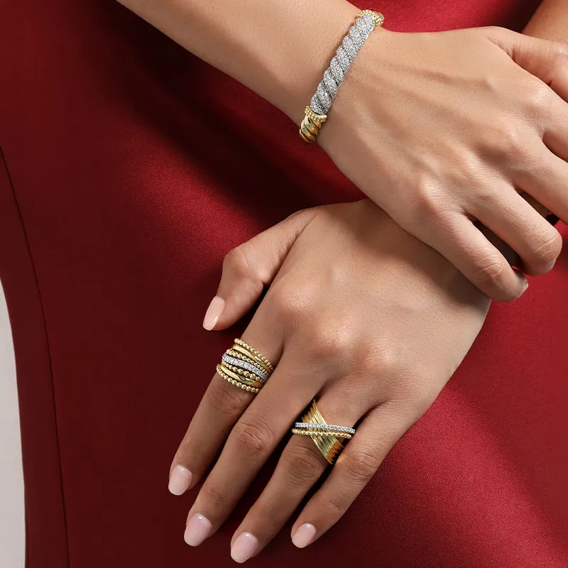 Hands adorned with gold and diamond rings and bracelet on a red dress background.
