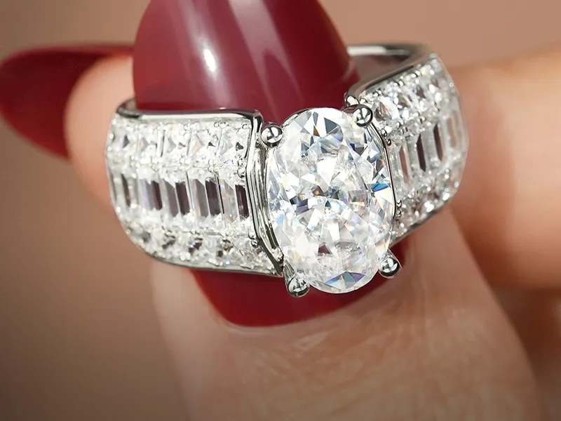 A silver ring with a large oval diamond and baguette accents held between fingers with red nails.