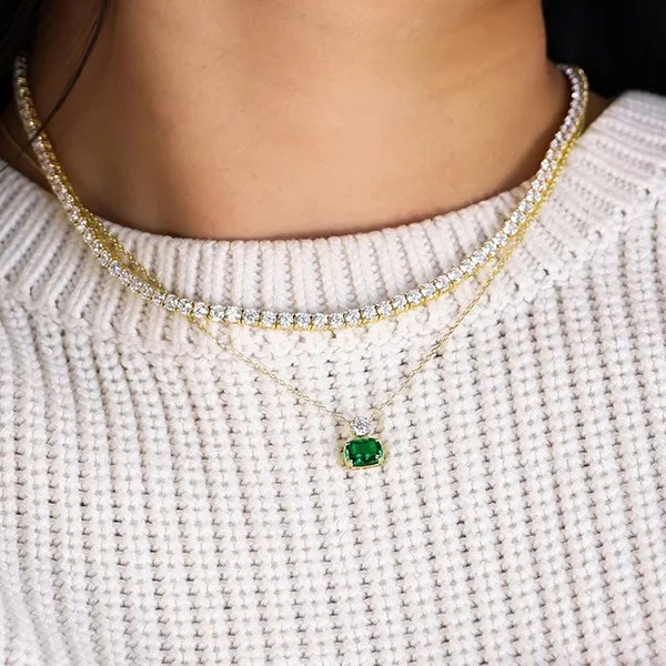 A person wearing layered necklaces with diamonds and a green gemstone pendant over a white sweater.